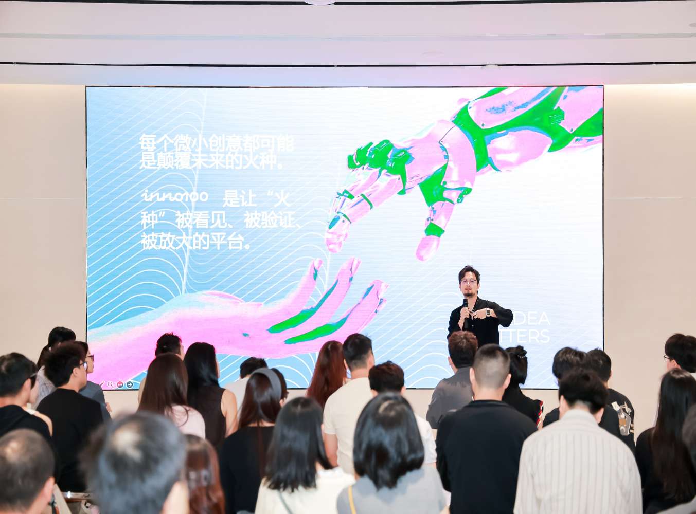 A speaker stands on stage addressing an audience in front of a large digital screen displaying artistic graphics and Chinese text at an innovation event